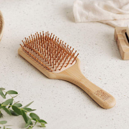 Jungle Culture organic bamboo paddle hairbrush with ergonomic handle and soft pins on a stone surface next to a green plant