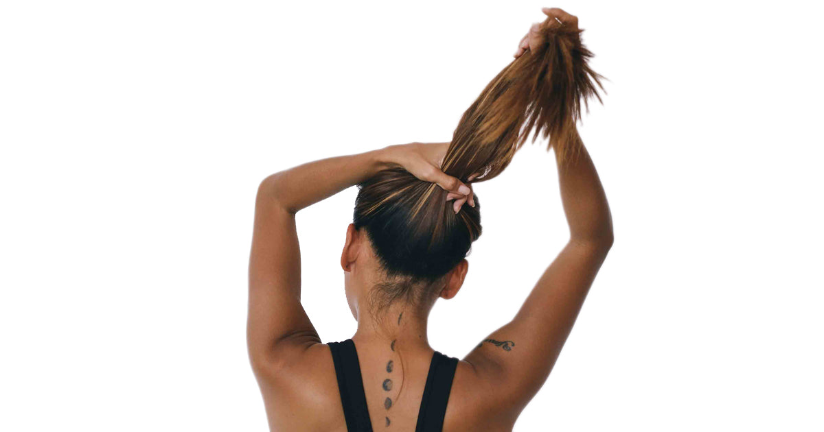 Back view of a woman with long hair and tattoos, pulling her hair up in a serene setting, representing Mybolife Self Care and Relaxation rituals.