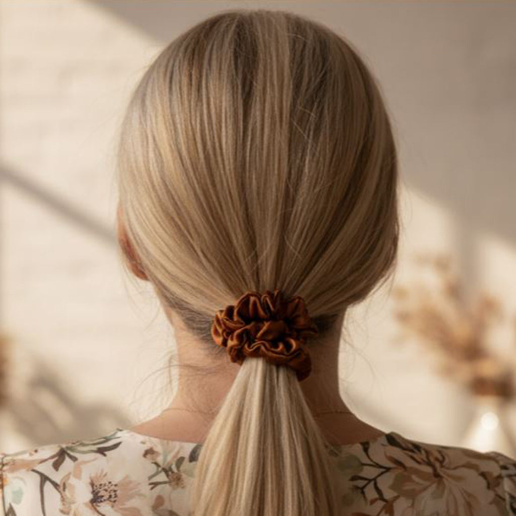 Person with blonde hair tied in a ponytail with a copper brown silk scrunchie, wearing a floral top.